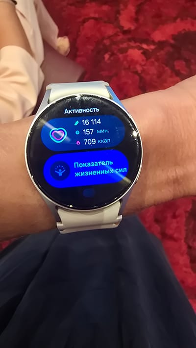 A close-up shot shows a person's wrist wearing a white smartwatch with a blue interface. The watch displays information related to activity. At the top, the word "Активность" (Activity) is visible. Below this, there are three lines of data: "16 114", "157 мин." (157 minutes), and "709 ккал" (709 kcal). A heart icon with a glowing outline is on the left side of the activity data. Further down the watch face, there is a blue section with an icon of a stylized person with outstretched arms and the text "Показатель жизненных сил" (Vitality indicator). The background is blurred, showing what appears to be a person in light-colored clothing and a textured red surface, possibly a sofa or carpet. The scene is likely indoors. There is no indication of the location being Agios Epiktitos, Northern Cyprus / Cyprus in the provided image.