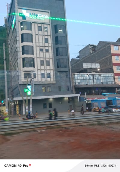 A tall, light-colored building labeled "Life Care Hospitals" dominates the left side. A green light strip runs across its top. The building stands adjacent to a road or train tracks, with several blurry figures and vehicles visible. Behind the hospital, a mix of older buildings is visible against a bright sky. The ground in the foreground is blurry and a reddish-brown color.