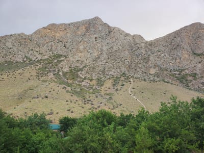 The image depicts a mountainous landscape with sparse vegetation in the foreground, composed of green trees and bushes. A small building with a green roof is partially visible amidst the foliage. The middle ground features a rocky, dry hillside with scattered shrubs and a visible hiking trail winding upwards. The background is dominated by a large, rugged mountain range characterized by its rocky texture and varying shades of gray and tan. The sky above is overcast and pale. The location is identified as Egizbulak, Uzbekistan. There are no people visible in the scene, nor any discernible actions or interactions.
