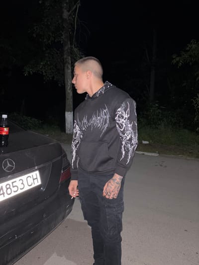 A person with a shaved head, wearing a dark hoodie with a white design and dark pants, stands next to a dark-colored car. The car's license plate reads "4853 CH". A bottle of Coca-Cola sits on the trunk of the car. The setting is a poorly lit area near some trees, possibly in a street. No information about the location can be determined beyond the car's license plate potentially indicating a location in Switzerland. No additional information regarding the city of Mihailovo, Bulgaria, can be determined from the image. The person is looking down at the car. The person is holding a cigarette in their hand.