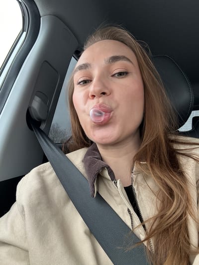 A young woman is seated in a car, wearing a light brown jacket and a seatbelt. She is blowing a bubble with gum. The interior of the car is visible, including the window, door panel, and black seats. The location of the scene is not identifiable from the image.