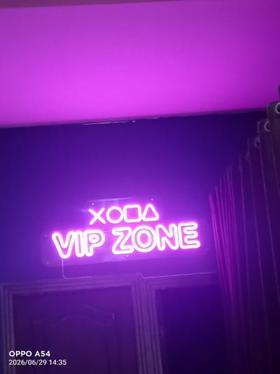 In Bokhtar, Tajikistan, a neon sign reads "VIP ZONE" in bold white lettering against a dark purple wall. Above the "VIP ZONE" text, a smaller neon sign displays a stylized symbol composed of a circle, a square, and a triangle. The lighting in the scene is predominantly purple, with a bright pinkish-purple illuminated ceiling. To the right, a dark curtain with vertical pleats is visible. At the bottom left of the image, a timestamp indicates "2026/06/29 14:35" and "OPPO A54". The scene appears to be an interior space, possibly a club or lounge, with a doorway visible below the neon sign.