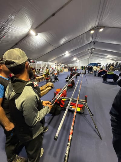 The image depicts an indoor exhibition or market within a large, white canvas tent structure, illuminated by overhead rectangular LED lights. The event is located in the city of Mora, Portugal, and features merchandise related to fishing. The floor is covered with a dark blue carpet.

In the foreground, a male individual is prominently featured from behind, wearing a light grey mesh trucker hat, a grey hooded sweatshirt over a black shirt, and dark pants. He has a dark beard and is actively examining a section of a fishing rod, holding it with both hands. To his right, two long fishing rods are displayed horizontally on black, tripod-style adjustable stands. The rods feature black base sections with red, white, and yellow graphics, including "BLACK SHADOW" and "CUSTOM RAGOT" text, and extend into silver-grey upper sections. Black bags and a red item are visible on the floor beneath the rods. A partially visible individual in dark clothing is present on the far right.

The midground and background reveal numerous people milling around various vendor stalls defined by white partition walls. These stalls display a variety of goods, including smaller items on tables, and larger equipment such as inflatable fishing vessels (float tubes) and other fishing gear. A blue umbrella covers one display area further back. The overall scene indicates an active trade show or fair focused on fishing equipment.