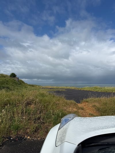 The image depicts a coastal landscape on an overcast day in Octeville-sur-Mer, France. The upper portion of the frame is dominated by a sky featuring extensive dark grey stratocumulus clouds, with interspersed patches of brighter blue sky visible in the top left quadrant. A distinct horizon line separates the sky from a calm, greenish-blue sea that occupies the middle ground.

To the left, a grassy hillside slopes downwards, covered in a mix of tall green and light brown dried grasses. Approximately halfway up the slope, a concrete structure, likely a bunker or coastal fortification, is partially visible, with a small, round green bush or tree growing on its upper surface.

In the mid-ground and extending towards the foreground, an irregularly shaped section of dark asphalt paving, resembling a disused road or viewing area, is present. Patches of red-brown dirt and green grass border and intrude upon this paved surface. The foreground on the right side displays the front-right quarter of a white vehicle, specifically its hood and part of the headlight assembly. The white hood is covered in numerous small water droplets, indicating recent precipitation. No individuals or active interactions are visible within the scene.