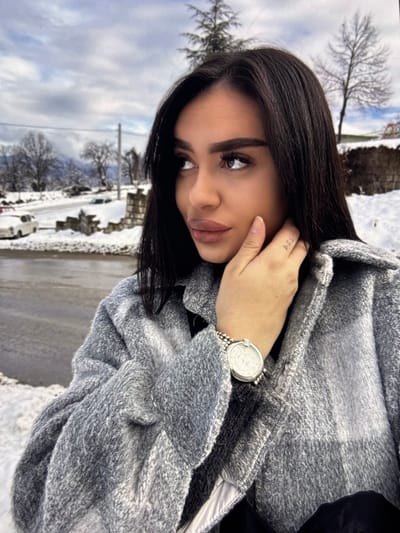 An outdoor, eye-level, slightly off-center portrait features a female individual against a snow-covered landscape in the city of Zhabokrat, Bulgaria. The subject, positioned in the right foreground, has long, dark, straight hair, prominent eyebrows, and full lips, looking upwards and to her right. Her right hand is raised, with fingers resting near her chin; a small, dark tattoo (possibly "A.C.J.") is visible on the lateral aspect of her right index finger. She wears a silver-toned watch with a round face and beaded strap on her left wrist. Her attire includes a grey, textured, possibly plaid, collared jacket over a dark garment.
The background depicts a winter scene with snow covering the ground, including a partially visible asphalt road to the left. Several bare deciduous trees and some evergreen foliage are present. A light-colored vehicle is parked on the snowy road shoulder to the far left. A low stone wall lines the road. Distant, snow-dusted hills or mountains are visible under an overcast, light grey sky.