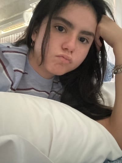 The image displays a young female, appearing to be in her late teens or early twenties, captured in a close-up, indoor setting. She possesses dark, long hair, dark eyes, and dark eyebrows. Her facial expression includes pouted lips and a subtle furrowed brow, with her direct gaze fixed on the camera. She is dressed in a light blue t-shirt with thin, horizontal maroon stripes. A gold-colored hoop earring is visible in her left ear, and her right wrist is adorned with multiple silver-colored link bracelets. Her right arm is positioned to support her head. She is lying on a white pillow that occupies the lower foreground of the image. The background is out of focus, showing a light-colored, wall-mounted air conditioning unit on the upper left and a blue patterned textile, possibly bedding or curtain, on the upper right. The environment suggests a bedroom or similar private accommodation. The specific location within the city of Santiago, Mexico, cannot be determined from the visual evidence presented in the image.