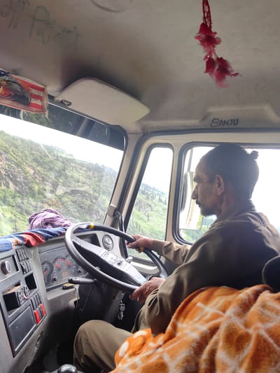 A man with a bun and a beard, wearing a brown shirt, is driving a truck. The truck's interior is visible, including the steering wheel, dashboard, and controls. An orange and white blanket is draped over the passenger seat. Outside the truck, a mountainous landscape with green vegetation is visible. The image is taken from inside the truck, looking towards the driver and the road ahead. The location is identified as Dulehar, India.
