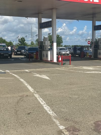 The image shows a gas station in Temryuk, Russia, with several cars lined up at the pumps. The sky is partly cloudy, and trees are visible in the background. There are also some power lines and utility poles. The gas station appears to be a typical one, with a canopy over the pumps and a small building for the cashier. The cars are various makes and models, and some people are visible standing around them. The overall scene is one of everyday activity at a gas station.
