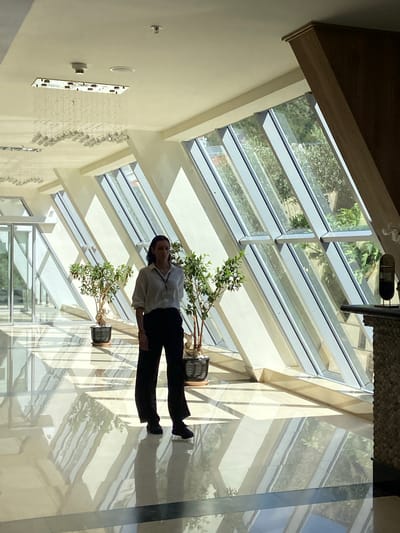 A woman stands in the middle of a modern building's interior hallway. The hallway has a high, angled ceiling with a decorative chandelier and a sprinkler system visible. Large, slanted windows line one side of the hallway, allowing sunlight to stream in and create geometric patterns on the polished floor. Two potted trees are placed strategically in the hallway. The woman is wearing a white collared shirt and black pants, with her dark hair down. She appears to be facing forward and is the sole human subject in the frame. The environment suggests an office building or a hotel lobby, and the lighting indicates it is daytime. The image does not provide any explicit information about the city of Brežine, Montenegro, beyond what is stated in the prompt.