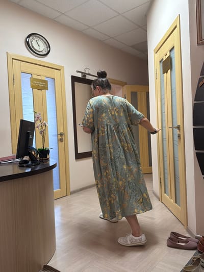 The image depicts an indoor scene, likely a hallway or reception area, based on the presence of doors, a reception desk, and a clock on the wall. A woman, seen from the back, is standing in the center of the frame. She has her hair pulled up in a bun and is wearing a long, light green dress with a floral pattern. She is holding a mobile phone in her right hand and appears to be walking towards a wooden door on the right side of the image. Her left arm is extended slightly to the side.
On the left side of the image, a curved reception desk is visible with a computer monitor and a potted orchid. Several doors are present, with signage on them, suggesting individual rooms or offices. A round clock with Roman numerals is mounted on the wall above one of the doors. The floor is a light-colored laminate with a wood grain pattern. In the foreground on the right, a pair of pink flat shoes are placed on the floor. The overall lighting is soft and ambient.
The signage on the doors is in Cyrillic script. One sign on a door on the left reads "КОЗБЕШКИН" and "ВАСИЛИЙ МАТВЕЕВИЧ", indicating a name and likely an office. Without additional context or clearer views of the signage, it's not possible to definitively place this location within Taganrog, Russia. However, the presence of Russian language signage strongly suggests a Russian context.