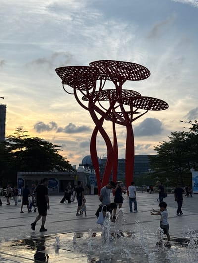 The image depicts a scene in the Nanshan District, China. It is an outdoor setting with a red, abstract, tree-like sculpture in the foreground. The sculpture consists of interconnected red metal pieces with circular, lattice-like shapes resembling the foliage of a tree.

Behind the sculpture, there is a view of a cityscape with buildings and structures, including the London Eye visible in the background. The sky is filled with clouds, reflecting colors of the setting sun, casting an orange and yellow glow.

On the right side of the image, there is a water feature, possibly a fountain or a shallow pool. People are interacting with the water, including a child playing with water. Other people are walking nearby on a paved surface. Trees line the edges of the paved area.