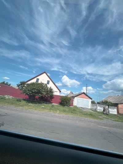 The image captures a rural or semi-rural residential street scene in Markivka, Ukraine, viewed from inside a vehicle, indicated by the dark car interior visible at the bottom. The sky, occupying the upper two-thirds of the frame, is clear blue with scattered white clouds, ranging from wispy cirrus-like streaks in the upper left to more defined cumulus clouds toward the right. Multiple thin power lines traverse the sky horizontally.

The foreground features a grey asphalt road, partially visible, with a grassy verge bordering the properties. No people are present.

On the left side of the midground, a two-story house with light-colored siding and a steep red gable roof is prominent. Two dark, rectangular windows are visible on its upper facade. A dense green deciduous tree stands directly in front of this house, partially obscuring its lower section. Behind the tree and house, a long red fence or wall extends horizontally.

To the right, further along the road, a brick outbuilding with a grey garage door and a dark roof is visible. A white, ornate fence with decorative balusters runs behind this building. A dark tree stump is situated near the fence. Another older structure with a dark, sloping roof and brick or earth-toned walls is partially visible beyond the fence on the far right. A wooden utility pole stands centrally in the mid-distance. Faint, bluish structures, possibly industrial or taller buildings, are discernible on the distant horizon to the right. The foliage is green, suggesting a spring or summer season under bright daylight.