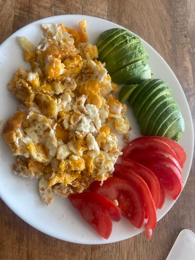 This is a top-down, close-up shot of a white plate filled with a breakfast meal, placed on a wooden table. The meal consists of scrambled eggs, sliced avocado, and sliced tomatoes. The scrambled eggs are in the foreground, appearing fluffy and slightly browned in places. To the left of the eggs, there is a fan of thinly sliced avocado, with its characteristic green skin visible on the outer edges. To the right of the avocado are several slices of bright red tomato, also fanned out.
The overall scene suggests a healthy and simple breakfast. The lighting is bright, indicating it could be daytime, likely morning, given the meal. There are no people or animals visible, and no text is present in the image. The wooden table has a visible grain texture. The white plate is clean and reflects some light. The arrangement of the food on the plate is neat, with distinct sections for each component of the meal. The close-up nature of the shot focuses the viewer's attention entirely on the food and the plate.