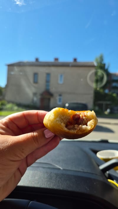 A close-up shot shows a hand holding a partially eaten pastry with a filling visible. The pastry is golden brown and appears to be soft. The hand has light skin with a pink nail polish on one fingernail. In the background, a light-colored building with a dark roof and multiple windows is out of focus. Trees and a clear blue sky are also visible in the background. The foreground shows the dark interior of a vehicle, possibly a car dashboard. A blurred white cup is also visible on the right side of the frame, indicating the scene is inside a vehicle. The setting suggests a casual moment, possibly a snack taken while traveling. The building in the background is a typical residential or small commercial building found in urban or suburban areas. The overall lighting indicates it is daytime with bright sunlight.