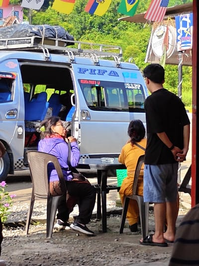In Tharpu, Nepal, at what appears to be a roadside stop, a silver van with "FIFA" and "BULLET" written on its side is parked with its doors open. Inside the van, blue seats are visible, and luggage is strapped to the roof rack. Two women are seated at a black table. One woman, wearing glasses and a purple hooded jacket over a patterned garment and black pants, gestures with her hands. The other woman, facing away from the camera, wears a bright yellow top and has her hair tied back. A young man, viewed from behind, stands nearby, wearing a black t-shirt and light blue shorts, with his hands clasped behind his back. A small table with an ashtray sits between the women. In the background, various national flags, including the American flag, are strung up, and a sign for "COFFEE" is visible. The ground is covered with gravel and small stones, and lush green foliage suggests a natural, outdoor setting.