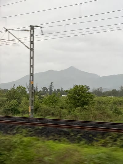 The image depicts a blurred, motion-effect view of a rural landscape in Kelva, India, likely taken from a moving train. A power line pylon with multiple horizontal wires dominates the left side of the frame, set against a hazy, overcast sky. In the middle ground, a dense band of lush green trees and foliage stretches horizontally across the image, with a hint of white structures peeking through in places. Further in the background, the soft, indistinct contours of misty mountains rise against the sky. In the foreground, blurred train tracks run horizontally, with a strip of blurred green vegetation below them, indicating the perspective of someone traveling by rail. The overall impression is one of a natural environment shrouded in a muted, atmospheric light.