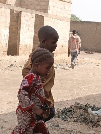 The image depicts an outdoor street scene in Zango, Nigeria, under bright daylight. In the foreground, two children are central subjects. An older child, with a dark complexion and short hair, wears a plain, light brown long-sleeved top and is positioned slightly behind and to the right, looking towards the right of the frame. A younger child, also with a dark complexion, has braided hair and wears a vibrant red and white patterned long dress. This child is in the immediate foreground, looking downwards and holding a dark, crumpled object in their hands.

In the mid-ground, an adult male walks away from the camera, towards the right. He wears a light-colored hooded sweatshirt with the distinct text "THE END" visible on the back, accompanied by a circular graphic (possibly a sun) next to "THE". He is also wearing light-colored jeans and appears to be holding a dark object in his right hand.

The setting is characterized by dusty, unpaved ground, indicative of a rural or less developed area. Piles of dark debris or waste are scattered, particularly noticeable on the lower right side of the image. A white chicken with some darker feathers is seen walking near one of these debris piles. The background features light-colored buildings constructed from blocks or bricks, some appearing unfinished or weathered, with rectangular openings that resemble doorways or windows. The overall scene suggests a casual, everyday moment in a village or semi-urban environment.