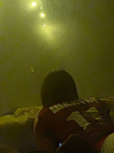 The image shows the back of a person wearing a red jersey with the number "1" on it. The person is lying down on a bed, with a textured green wall behind them. There are small, bright lights strung along the wall, casting a soft glow. The overall scene appears to be indoors, likely a bedroom. There is no identifiable information about the location in the city of Shakhty, Russia.