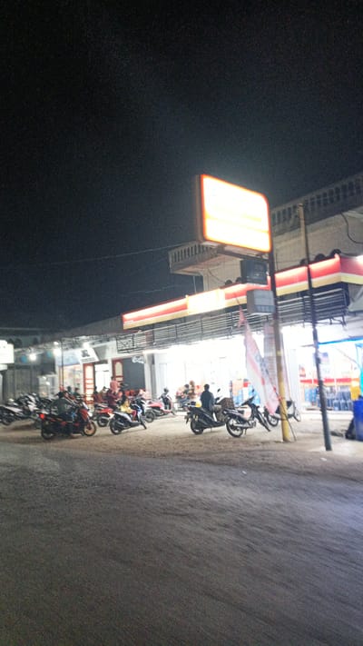This image captures a nighttime scene outside a brightly lit convenience store in Uteungeulinggang, Indonesia. The store, identified by its red and yellow signage and awning, appears to be open, with lights illuminating its interior and exterior. A prominent illuminated sign, likely for the store, hangs high above the entrance. In front of the store, a paved area is filled with numerous parked motorcycles, suggesting it's a popular spot. Several individuals are visible, some standing near their motorcycles, while others are seated on them, indicating people are either arriving, departing, or taking a break. The ground surface is rough and unpaved in the foreground, transitioning to a smoother paved area near the store and motorcycles. The overall atmosphere is one of a bustling local hub at night.