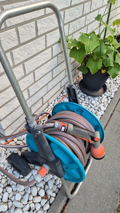 The image captures a close-up view of a garden hose reel with a coiled hose, a watering nozzle, and a small wheel, situated on a bed of white pebbles next to a white brick wall and a concrete walkway. In the background, a potted cucumber plant with green leaves and a yellow flower is visible. The scene appears to be outdoors, likely in a residential garden, and is consistent with locations in Dissen am Teutoburger Wald, Germany, known for its gardening and outdoor spaces. No people are present in the image, and the objects are stationary. The lighting suggests it is daytime, with natural light illuminating the scene.
