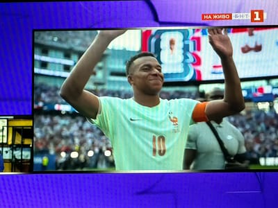 A soccer player, wearing a light green jersey with the number "10" on the front and an orange armband, is shown on a television screen. The player has dark hair and is smiling, with his right arm raised. In the background, a crowd of spectators is visible, along with a scoreboard or graphic displaying "НА ЖИВО Б Н Т 1". The image appears to be a screenshot of a broadcast, possibly from a sporting event. The television is displayed on a large, purple screen or panel, suggesting it's part of a larger display or setup, potentially in Krumovgrad, Bulgaria, though this cannot be confirmed from the image itself.