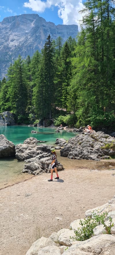 The image captures a scenic view of a turquoise lake in a mountainous region, likely the Dolomites in Italy given the context of Pragser Wildsee (Lago di Braies). The scene is oriented vertically, with the lake and its rocky shore in the foreground and a dense forest of evergreen trees and a towering mountain in the background. The water is remarkably clear, revealing the rocky bottom.

Several people are visible along the shore. In the foreground, a person wearing a cycling outfit with a helmet stands on the gravelly bank next to the water. Further back, another person in a red shirt and white shorts is crouched on a rock by the water's edge. A third person is visible in the water, near the edge, possibly swimming or wading.

The time of day appears to be daytime, indicated by the bright sunlight casting shadows. The weather seems pleasant, with clear blue skies and scattered clouds visible behind the mountain. The overall atmosphere is serene and recreational, suggesting a popular spot for outdoor activities like hiking or cycling, with the vibrant colors of nature dominating the scene. There is no visible text in the image.
