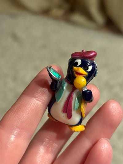 A colorful, decorative penguin figurine is held in a person's fingers. The penguin has a white belly, dark blue head and body, and a red hat. A yellow bow tie is attached to its neck. There are colored ribbons or accents in shades of teal and maroon on its chest. The penguin's beak is yellow, and one foot has a shimmery, iridescent finish. The background is blurry but appears to be a neutral color.