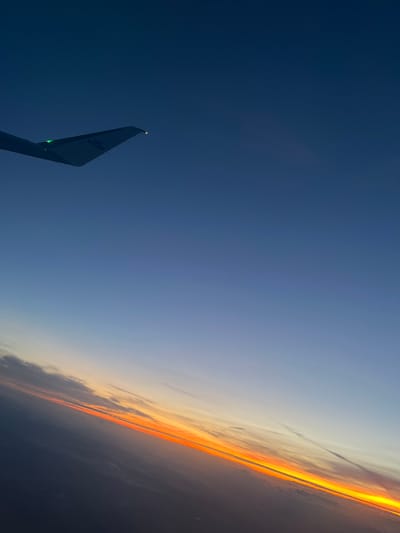 The image shows a view from an airplane window at sunset. The wing of a KLM plane is visible in the lower left corner. Above the wing, the sky transitions from a dark blue to a light blue. On the right side of the image, the horizon is visible with a bright orange and yellow sunset. The city of Ettelbruck, Luxembourg is not visible in the image.