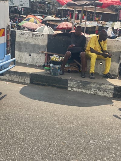 The image depicts a bustling urban scene in Awjaw, Nigeria, centered on two adult males seated alongside a road. In the foreground, a dark asphalt road surface is visible, leading to a concrete curb painted with white and black stripes. Elevated behind the curb is a paved area with two men seated on a low wooden bench positioned against a two-tiered concrete barrier, also painted white and black.
The man on the left has a dark complexion, is bald, and wears a dark long-sleeved shirt, dark shorts, and dark shoes. He rests his left arm on the top of the concrete barrier and wears a watch on his left wrist. The man on the right, also with a dark complexion and bald, wears a bright yellow long-sleeved shirt, yellow trousers, and dark sandals. His hands are clasped in front of him.
Between the two men, and in front of the left man, several items are arranged on the ground and on a wooden support below the bench. These include a black bowl filled with individually packaged items, and various plastic containers—a green bucket, a clear bucket, and a white bucket—some containing merchandise. A brown woven basket is visible beneath the bench.
To the far left, the light blue and white rear-side of a vehicle, possibly a bus, is partially visible, displaying red and yellow hazard stripes. In the midground, behind the concrete barrier and the men, a vibrant street market extends. Numerous market stalls are visible, characterized by varied colorful umbrellas (red, white, pink, green) providing shade, and corrugated metal roofing. People are actively moving and congregating within the market area. The background features indistinct structures, likely market buildings, under a bright, clear sky. Strong shadows are cast on the asphalt road, indicating bright daylight conditions.