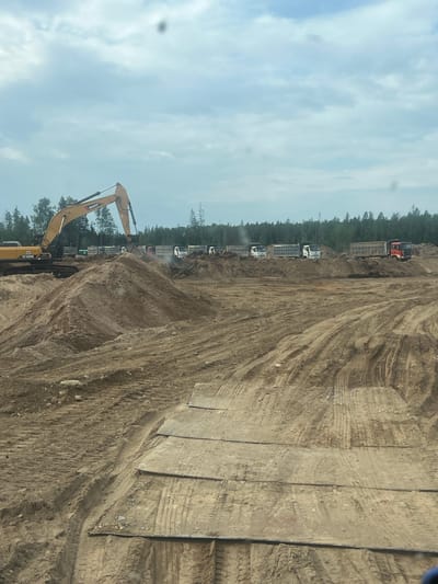 The image depicts a construction site in Shapki, Russia, on a cloudy day. A large yellow excavator is actively digging into a mound of dirt, with its bucket full. In the background, a line of dump trucks is parked, suggesting a large-scale earthmoving operation. The terrain is uneven and appears to be undergoing significant excavation and grading, with tire tracks visible in the dirt. The surrounding area is bordered by a dense line of trees.