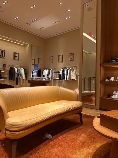 The scene depicts the interior of an upscale retail establishment located in Winchester, United States of America. The environment is brightly illuminated and currently devoid of human presence.

In the foreground, a long, curved sofa upholstered in light tan leather with dark wooden legs rests on an orange-brown shag carpet. A crumpled piece of light-colored plastic or paper is visible on the carpet beneath the sofa. To the right, a section of a circular wooden coffee table is visible, supporting a rectangular wooden box.

The midground features light beige walls adorned with multiple framed abstract artworks. Two wall-mounted metal racks display folded and hanging garments, including shirts and light jackets, primarily in white, grey, blue, and black tones. A central glass-topped display case on a wooden pedestal is present. A large, full-length mirror on the right wall reflects an adjacent hallway or stairwell with wooden railings and a partial red "EXIT" sign overhead.

To the far right, a tall wooden shelving unit contains multiple pairs of shoes, including dark loafers and various sneakers, displayed on its tiers. The light-colored ceiling is equipped with numerous recessed circular spotlights that provide uniform illumination to the space.