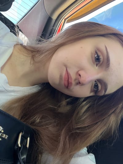 A close-up, low-angle shot shows a young woman with light brown hair tilted her head, looking towards the camera. She is wearing a white t-shirt and a black purse with gold hardware is visible in the foreground. The background appears to be the interior of a car, with sunlight filtering through the rear window. The image does not contain any information about the city of Марьевка, Russia.