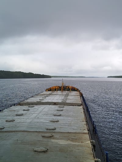 The image shows a view from the deck of a ship moving through a body of water, likely a wide river or lake. The forward deck of the ship, consisting of gray panels with circular access hatches, stretches into the distance. The sides of the ship are visible, with a blue railing along the edge. The water is choppy with a slight ripple texture. On either side of the waterway, land is visible, densely covered with green trees. The sky is overcast with thick gray clouds. The scene suggests a cloudy or rainy day. There is no indication of this scene being near Irma, Russia.