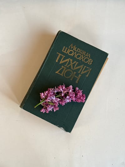 A slightly angled top-down shot displays a green hardback book with Cyrillic text in gold lettering. A sprig of lilac flowers rests on top of the book. The background is a plain, off-white surface. There are no people in the shot. The book's title is "Quiet Flows the Don" by Mikhail Sholokhov. The location, Ленино, Russia, is not identifiable from the image.