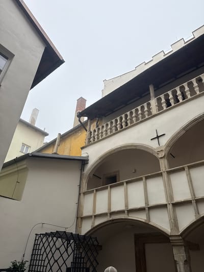 The image displays an architectural scene within a courtyard or narrow passage between buildings in the city of Znojmo, Czechia, under an overcast sky. The dominant structure on the right is a multi-story building constructed primarily of light-colored stone or stucco. The uppermost visible section features a dark wooden eaves or balcony roof, beneath which is a stone balustrade with ornate balusters. Above this, a crenellated parapet is visible against the grey sky. Below the balustrade, an arcade level is present, characterized by a series of rounded stone arches forming an open loggia. A dark, possibly metal, cross-shaped fixture is affixed to the wall between two arches. Below this arcade, a second, lower balcony or walkway is visible, featuring a robust, light-colored stone balustrade composed of rectangular panels and vertical supports, which are in turn supported by further arches beneath.

To the left, a plainer, light-beige building with a sloped, dark-tiled roof and a single rectangular window on an upper floor is partially visible. Further in the background, a building with a weathered yellow-ochre facade and a red brick chimney rises above the adjacent structures. Another tall, slender, light-colored chimney is visible in the more distant background. In the lower-left foreground, a dark, diamond-patterned metal structure, possibly a trellis, is partially visible, with a small amount of green foliage at its base. A light-colored, rounded object, potentially a decorative cap or base, is partially visible at the very bottom center of the frame. No individuals are present in the scene. The overall lighting is soft and diffused, indicative of heavy cloud cover.