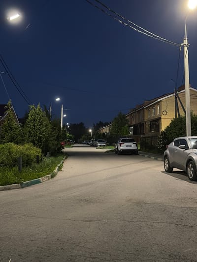 The image displays a residential street in Новинки, Russia, observed at night or during twilight, indicated by the dark blue sky and active artificial lighting. A paved asphalt road, showing signs of wear and faint tire marks, occupies the center and foreground. A concrete curb, painted with light green and white sections, delineates the road's left edge, adjacent to an area of grass and low shrubs.
On the right side of the street, a silver compact SUV is partially visible in the immediate foreground. Further along the right, a white Geely Coolray SUV, bearing Russian license plate A927CX, is parked. A series of two-story yellow brick townhouses with brown roofs line the right side of the street, separated from the road by a dark metal fence. One townhouse features a dark, ring-shaped object on its exterior wall. Multiple streetlights, emitting a bright, yellowish-white light, illuminate the street and buildings. Overhead utility lines are present across the upper portion of the frame.
On the left side of the street, a dense arrangement of coniferous trees and other vegetation screens a house with a red roof, which is partially visible. Additional parked vehicles are discernible further down the street on both sides, progressively less distinct due to distance. No individuals are present in the scene.