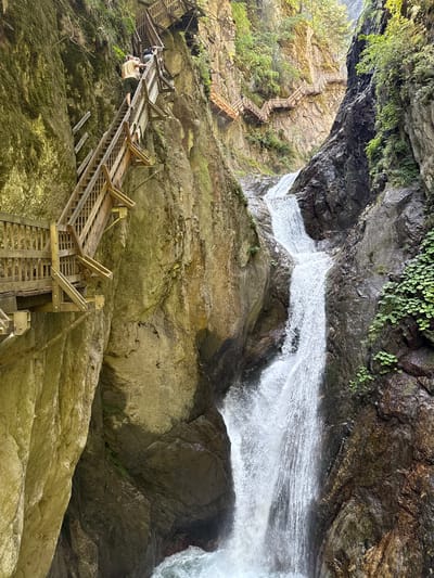 The image depicts the Gorge du Trient, a spectacular natural canyon located in the municipality of Trient, within the canton of Valais, Switzerland, near Martigny-Combe. The scene is dominated by a cascading waterfall plunging into a rocky gorge. Wooden walkways and staircases are constructed along the steep canyon walls, allowing visitors to navigate the dramatic terrain and experience the waterfall up close. In the foreground, two individuals are ascending one of the wooden staircases, their figures dwarfed by the sheer scale of the natural environment and the man-made structures. Vegetation, including moss and small plants, clings to the rocky surfaces. The sunlight filters through the gorge, illuminating parts of the rock faces and the mist rising from the waterfall.