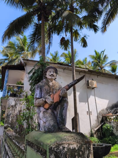 A weathered statue of a man in a hat and coat, playing a stringed instrument, kneels on a moss-covered pedestal. The statue is the primary subject, located in the foreground, and is a focal point of the image. Behind the statue, a white building with a corrugated metal roof is visible, featuring an air conditioning unit mounted on the exterior wall. Palm trees, with lush green fronds, dominate the background, indicating a tropical or subtropical environment. The scene is outdoors, under a clear blue sky. The overall impression is one of a weathered, possibly aged, outdoor setting with a rustic statue as a central element. The location is identified as Oxel, India.