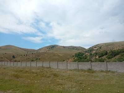 The image depicts a rural landscape under a cloudy sky. In the foreground, a long, low wall constructed from grey concrete blocks stretches horizontally across the frame, supported by vertical posts. Beyond the wall, a grassy field with scattered yellow wildflowers is visible. The midground is dominated by rolling hills covered in dry, golden-brown vegetation and sparse green trees. A narrow road with a red-tiled roof structure is partially visible behind the wall on the left. The background features more undulating hills, some with rocky outcrops and patches of greenery. The overall scene suggests a dry, open environment with a man-made barrier separating the immediate foreground from the natural landscape. This scene is located in the vicinity of Көксаяқ ауылдық округі, Kazakhstan.