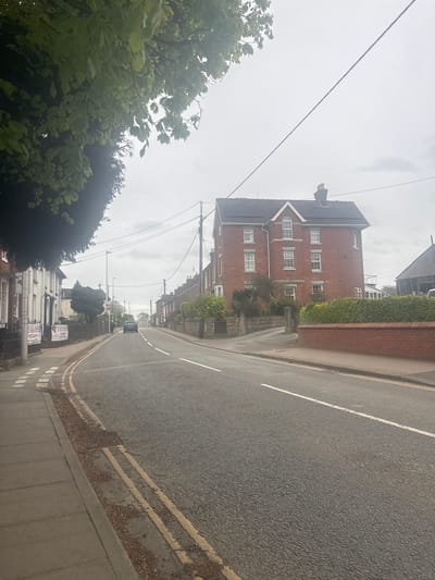 This image depicts a quiet street scene in Welshpool, United Kingdom, under an overcast sky. The photograph is taken from a tilted perspective, showing the street ascending diagonally from bottom right to top left.
On the right side, an asphalt road features clear white dashed lines and double yellow no-parking lines along the curb. A grey pavement runs parallel to the road, bordered by a low brick wall adorned with lush green ivy or shrubbery. Behind this, a substantial red brick building stands prominently, characterized by multiple white-framed windows, including an arched window on a gable, and a dark, possibly tiled roof with some dark panels (potentially solar panels).
The upper part of the frame is dominated by a pale, cloudy sky crisscrossed by several overhead utility lines and poles. In the lower left, dense green foliage from trees partially obscures the view of the street and buildings beyond. Further down the street on the left, a row of residential buildings, some painted white, are visible. A dark-colored car is parked further in the distance on the left side of the road.
Visible text on signs along the left pavement includes "THE ORCHID" and directly below it, "PARAYAN'S." The overall weather appears dull and grey, indicative of an overcast daytime. There are no people visible, and no specific activity is taking place, presenting a serene, almost deserted urban landscape.