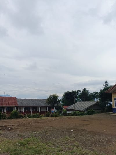 The image depicts an outdoor scene in São Félix do Xingu, Brazil, under a heavily overcast sky composed of light gray clouds. A small, dark bird is visible in flight in the upper left quadrant. The foreground features an expansive, uneven ground composed primarily of packed dirt and sparse, dry grass with scattered small debris. In the midground, a series of structures are visible. To the left, a long, single-story building with a red-tiled roof section on the far left transitions to a dark gray roof, featuring a white facade with red trim and multiple rectangular windows. Further right, partially obscured by dense green foliage, is another single-story building with a dark gray, sloped roof. A portion of a third building, characterized by a light yellow wall, a blue base, and a red-tiled roof, is visible on the far right. Lush green trees and shrubs are interspersed among and behind the structures. Distant, hazy mountains or hills are visible on the horizon line in the background to the left. No individuals, actions, or interactions are observed within the scene.