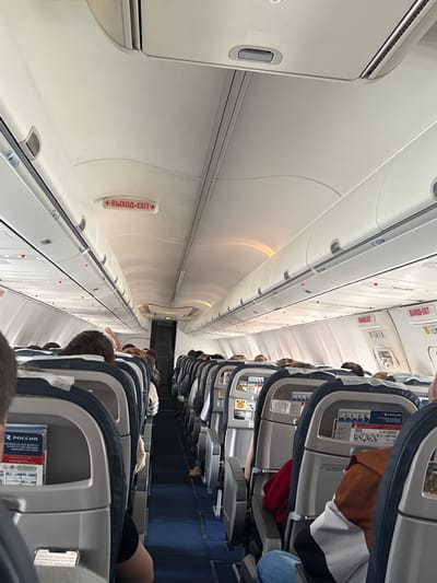 The scene depicts the interior of a commercial passenger aircraft cabin, identified as a Boeing 737-800 operated by Rossiya Airlines, presumably associated with operations at Ufa Airport, Russia. The view is from a seated passenger's perspective, looking towards the rear of the aircraft down a central blue-carpeted aisle. Overhead white paneling and closed luggage compartments line both sides of the cabin. Multiple illuminated red "ВЫХОД-EXIT" signs are visible along the overhead panels, some with directional arrows. Numerous passengers are seated in the grey and blue upholstered seats, predominantly facing forward. In the immediate foreground, a smartphone is partially visible in a seatback pocket on the left. Further down the left aisle, a passenger is seen with an arm raised. Safety information cards bearing the "РОССИЯ" logo are present in the seatback pockets of several seats. Artificial interior lighting illuminates the cabin.