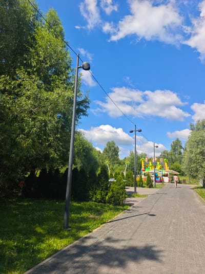 The image depicts a paved walkway in a park on a sunny day. Tall green trees line the left side of the walkway, with a grassy area in front of them. Several lampposts are visible along the path. In the distance, a colorful inflatable playground structure with cartoon animals is set up, suggesting it's a recreational area. A lone person, a woman wearing a light brown outfit, is walking away from the camera on the walkway, heading towards the playground. The sky is bright blue with scattered white clouds. The scene appears to be in a park or amusement area within the city of Chekhov, Russia, given the context of the location provided.
