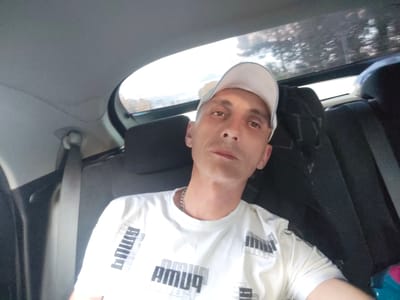 In this close-up, self-taken photograph, a Caucasian man with short, light-colored hair and a light-colored baseball cap is seated in the back seat of a car. He is wearing a white t-shirt with a repeating black and white "PUMA" logo design. He has a silver chain around his neck. The background shows the interior of a car, including the headrests of the seats and a portion of the rear window, through which trees and a bright sky are visible. The image appears to have been taken during daylight. There is no specific visual information within the image to determine the exact location within the city of Bor, Russia.