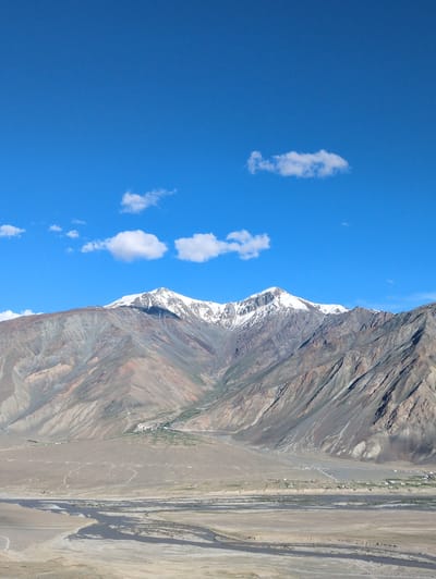 The image depicts a panoramic view of the mountains near Yulang, India. The sky is a vibrant blue, dotted with a few white clouds. In the foreground, a wide, meandering river flows through a dry, sandy landscape. The riverbed shows signs of varying water levels, with exposed sandbanks and darker, deeper channels.
Further back, the landscape transitions to a flatter plain where a small village is nestled at the foot of the mountains. The village appears to consist of clusters of white buildings, surrounded by patches of green vegetation, suggesting agricultural activity.
Dominating the background are the majestic mountains. Their slopes are a mosaic of earthy tones, ranging from sandy browns and muted grays to streaks of reddish-orange. The upper reaches of the mountains are capped with snow, creating a stark contrast against the clear blue sky. The mountain faces are deeply carved by ravines and geological formations, indicative of a rugged and ancient terrain. The overall scene conveys a sense of vastness and natural grandeur.