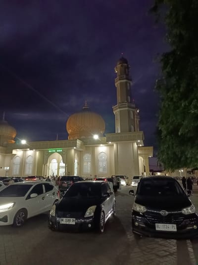 The image depicts the Baitur Rrahman Grand Mosque in Mee Tanjong Usi, Indonesia, at night. The mosque features a large golden dome and a tall minaret, illuminated by exterior lights. In the foreground, a parking lot is filled with several cars, including a white hatchback on the left, a black hatchback in the center with license plate "BK 1088 OH", and a black SUV on the right with license plate "BL 1724 PL". People are visible walking around the mosque entrance and in the parking area. The sky is dark blue with some clouds. Lush green trees frame the right side of the image.