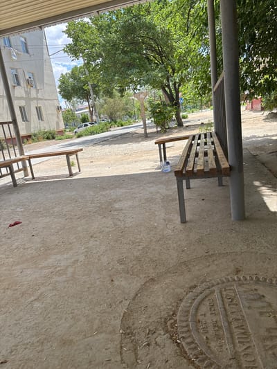 The image depicts an outdoor scene in Nukus, Uzbekistan, likely during daylight hours. The perspective is low and angled, giving a slightly disorienting view. In the foreground, a simple wooden bench with metal legs rests on a concrete surface. A clear plastic bottle of water sits on the bench. To the left, a metal support pole and ribbed metal roofing suggest a covered walkway or structure.

Behind the bench, a paved area stretches into the background, lined with sparse greenery and trees. Further back, a white apartment building with visible air conditioning units and windows is present. A white car is parked on the side of the road. The sky is partly cloudy.

A circular manhole cover is embedded in the concrete to the right of the bench. Visible text on the cover includes "ГОСТ 3634" and "T 250," indicating a standard and possibly a size or weight rating. Another section of text, partially obscured, appears to read "УЗКРАЙКОМ..." which might refer to a regional committee or organization. The scene conveys a sense of everyday urban life, with a focus on functional infrastructure and simple public amenities. The overall impression is one of a warm, possibly summer day.