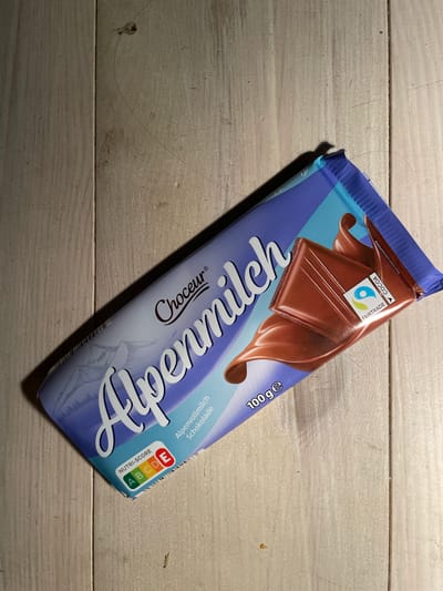 A chocolate bar with the branding "Choceur Alpenmilch" is placed diagonally on a light-colored wooden surface. The chocolate bar is partially unwrapped, revealing a section of the chocolate. The packaging is blue and white with mountain imagery. There are several logos on the packaging, including "Nutri-Score" with a rating of C and "Fairtrade." The scene is lit from the side, casting a shadow to the left of the chocolate bar. The wooden surface appears to be made of planks and shows some texture and wear. There are no people or animals visible in the image. The image does not provide any information about the location in the city of Oldenburg in Holstein, Germany.
