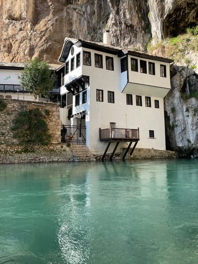 This image displays a multi-story white building with prominent dark wooden trim and window frames, situated directly against a large, steep rock cliff face. The structure is located in Dračevice, Bosnia and Herzegovina. A wide, tranquil body of turquoise-colored water, likely a river, occupies the foreground, reflecting portions of the building and the sky.
The building features a distinctive dark, curved roofline and multiple levels with numerous windows. A wooden balcony with vertical railings extends over the water on the building's right side, supported by two angled dark beams. To the left of the main structure, a stone retaining wall supports a small green tree, and a section of a white wall bears green stylized script. At the base of the building, on the left, stone steps lead down to a dark metal gate.
Two individuals are visible near the gate: an adult figure in dark clothing is partially obscured behind it, while a child in a red top stands in front of the gate with arms extended horizontally. Higher on the building, a human head and shoulders are discernible through an upper-story window on the right facade, oriented outwards. The water surface exhibits slight ripples and distorted reflections of the surrounding environment.