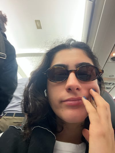 The image shows a close-up selfie of a young woman wearing sunglasses and a black jacket. She is indoors, in what appears to be a plane cabin, with a light-colored ceiling and overhead compartments visible. The woman's left hand is touching her cheek. The background includes a portion of another person's back and shoulder, also appearing to be on the plane. The lighting is bright and appears to be artificial.